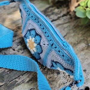 May include: A handmade crochet crossbody bag in shades of blue, gray, and white. The bag features a floral design and a long, adjustable strap. The bag has a zipper closure and metal hardware.