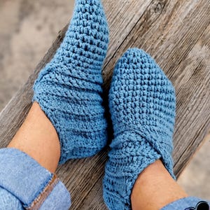 Puede incluir: Un par de zapatillas de ganchillo azules hechas a mano. Las zapatillas son de color azul claro liso y parecen estar hechas con un punto texturizado. Las zapatillas se muestran sobre una superficie de madera.
