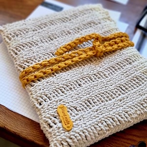 Puede incluir: Una funda para portátil beige tejida a ganchillo con un cierre de lazo amarillo mostaza. La funda tiene un patrón texturizado y una pequeña etiqueta marrón que dice "Handmade with love".