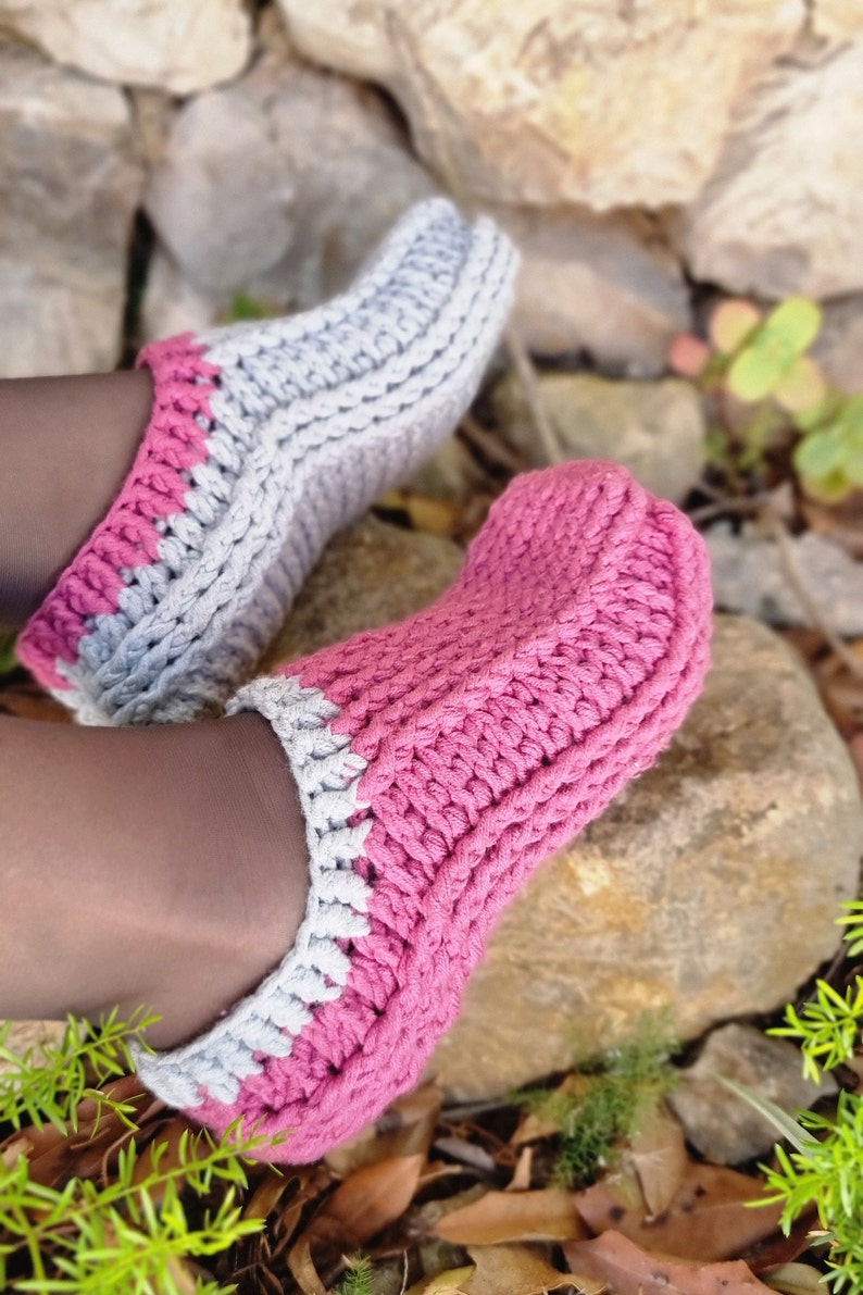 May include: A pair of handmade crochet slippers in shades of pink and gray. The slippers have a thick, textured stitch pattern and a contrasting color trim. The cozy footwear is perfect for indoor wear.