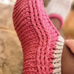 May include: Close-up of a handmade, pink and gray knitted slipper. The slipper is thick and textured, with a ribbed design. The toe and heel are a solid pink, while the cuff is gray with a pink trim. The slipper is designed for comfort and warmth.