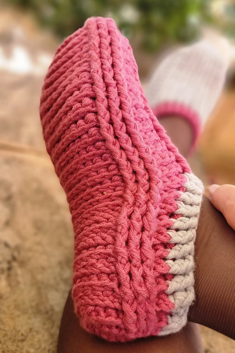 May include: Close-up of a handmade, pink and gray knitted slipper. The slipper is thick and textured, with a ribbed design. The toe and heel are a solid pink, while the cuff is gray with a pink trim. The slipper is designed for comfort and warmth.