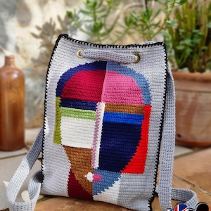 May include: A grey crochet backpack featuring a colourful abstract face design. The face is made up of red, pink, blue, green, and brown geometric shapes. The bag has a black border, grey straps, and gold-coloured metal eyelets.