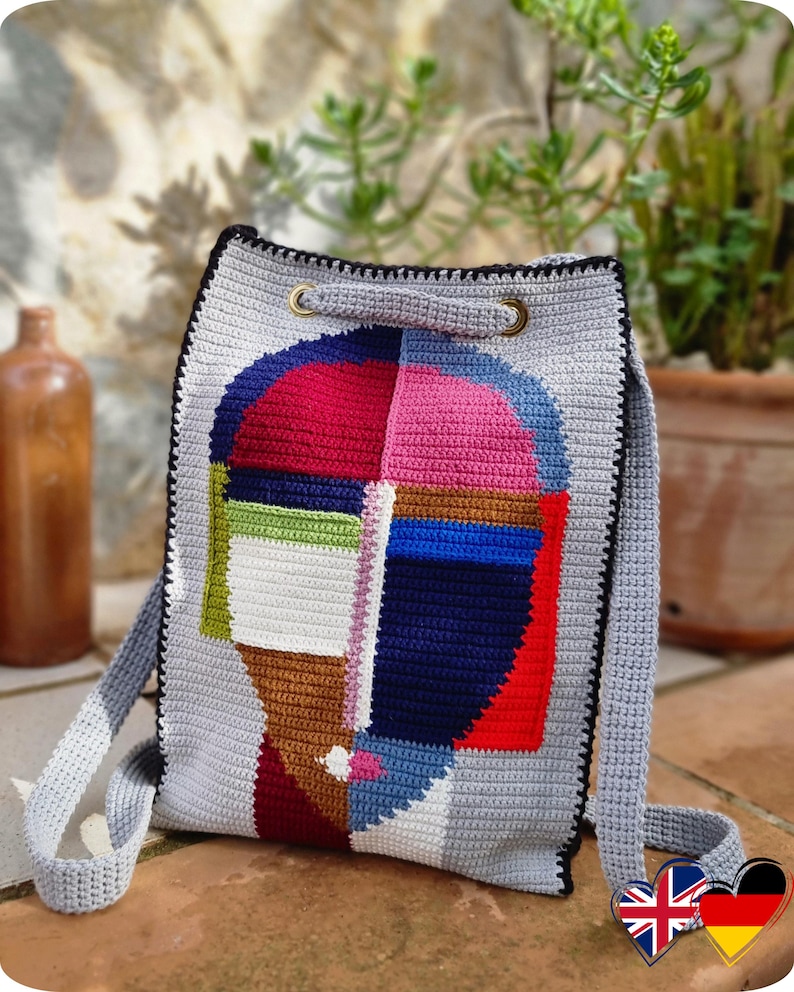 May include: A gray crochet backpack with a colorful abstract face design. The face is composed of red, pink, blue, green, and brown geometric shapes. The bag has a black border, gray straps, and gold-colored metal eyelets.