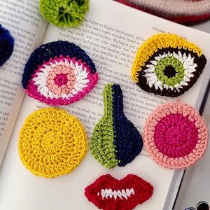 Puede incluir: Una colección de apliques de ganchillo coloridos en varias formas, incluyendo un ojo, labios y un círculo, colocados sobre un libro abierto. Los apliques presentan colores brillantes como amarillo, rosa y verde, y están hechos a mano.