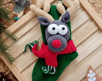 May include: A green crochet Christmas stocking featuring a three-dimensional gray reindeer head with large white eyes, a red nose, and beige antlers. The reindeer wears a bright red crochet scarf with green and white fringe accents.