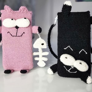 Op de afbeelding: Twee gehaakte telefoonhoesjes in de vorm van een kat, &eacute;&eacute;n roze en &eacute;&eacute;n zwart. De roze hoes heeft een witte band en houdt een witte visgraatbedel vast. De zwarte hoes heeft een zwarte band. Beide hoesjes hebben witte ogen en poten.