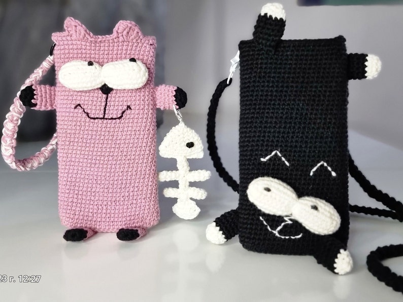 Op de afbeelding: Twee gehaakte telefoonhoesjes in de vorm van een kat, &eacute;&eacute;n roze en &eacute;&eacute;n zwart. De roze hoes heeft een witte band en houdt een witte visgraatbedel vast. De zwarte hoes heeft een zwarte band. Beide hoesjes hebben witte ogen en poten.