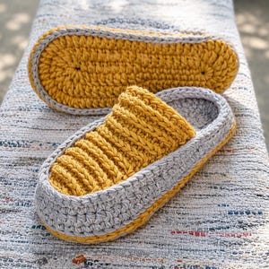 May include: Handmade crochet slippers in mustard yellow and gray. The slippers have a ribbed design on the top and a solid sole. The second slipper is placed next to the first one. The slippers are on a textured surface.