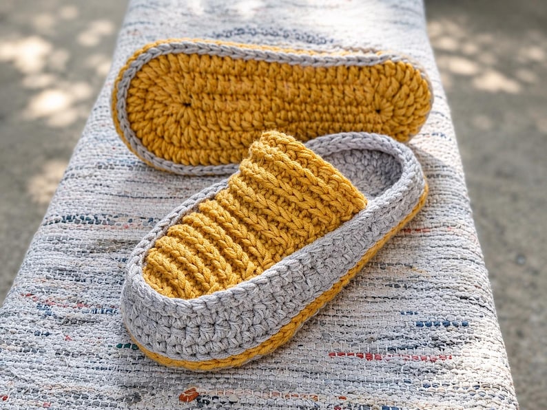 May include: Handmade crochet slippers in mustard yellow and gray. The slippers have a ribbed design on the top and a solid sole. The second slipper is placed next to the first one. The slippers are on a textured surface.