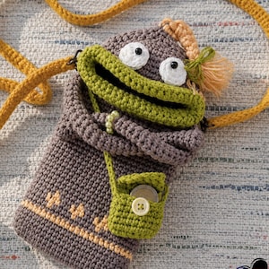 Può includere: Borsa a tracolla all'uncinetto fatta a mano a forma di creatura stravagante. La borsa è principalmente grigia con una bocca verde, occhi bianchi e un piccolo portamonete verde. Ha una tracolla color senape.