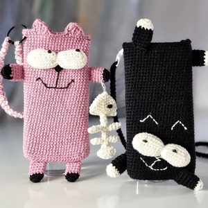 Op de afbeelding: Twee gehaakte telefoonhoesjes in de vorm van een kat. E&eacute;n is roze met zwarte en witte accenten, de andere is zwart met witte accenten. Beide hebben een lachend gezichtsontwerp en een riem. De roze kat heeft een visgraatbedel.