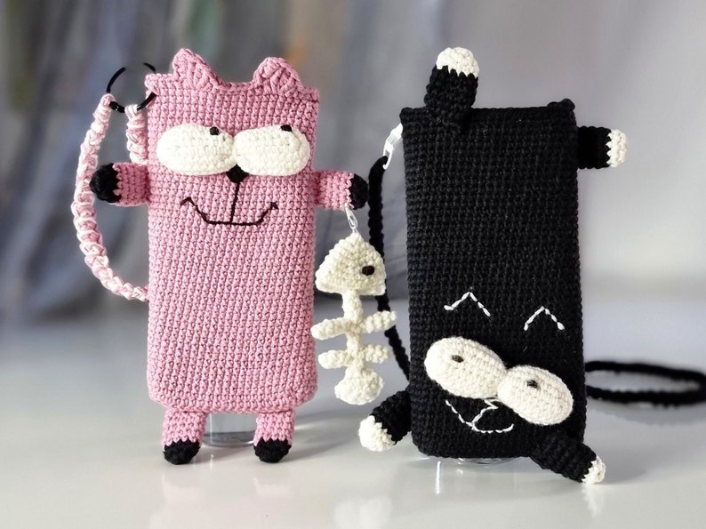 Op de afbeelding: Twee gehaakte telefoonhoesjes in de vorm van een kat. E&eacute;n is roze met zwarte en witte accenten, de andere is zwart met witte accenten. Beide hebben een lachend gezichtsontwerp en een riem. De roze kat heeft een visgraatbedel.