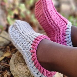 May include: A pair of handmade crochet slippers. The slippers are gray with a pink trim around the opening and on the top of the foot. The slippers are resting on a rock, with a blurred background of greenery.