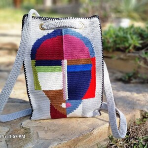 May include: A handmade crochet backpack with a geometric face design. The bag features a gray body with a colorful face design in red, blue, pink, green, and brown. It has gray straps and black trim, with gold-colored metal rings at the top.