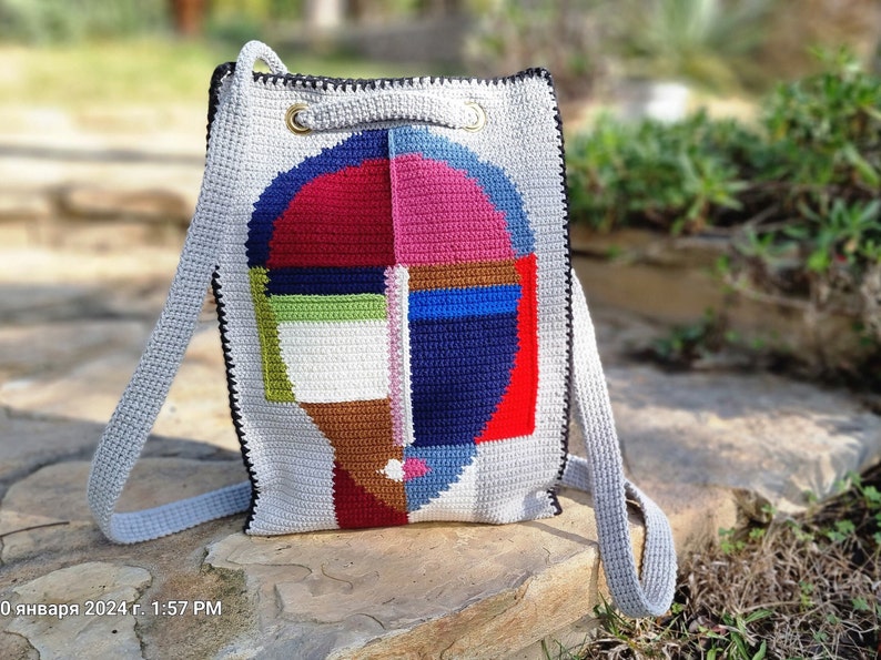 May include: A handmade crochet backpack with a geometric face design. The bag features a gray body with a colorful face design in red, blue, pink, green, and brown. It has gray straps and black trim, with gold-colored metal rings at the top.