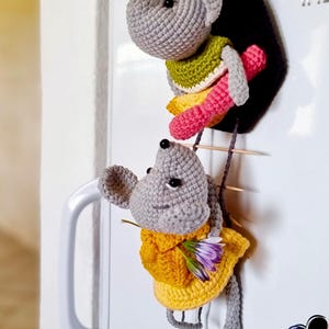 Può includere: Due figure di topi all'uncinetto fatte a mano fissate su una superficie bianca. Il topo in alto è grigio con un top verde e zampe rosa. Il topo in basso è grigio con un vestito giallo e tiene in mano dei piccoli fiori viola.