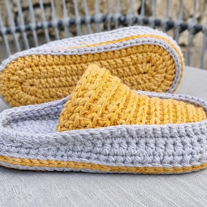 May include: Handmade crochet slippers in gray and yellow. The slippers feature a gray base with a yellow sole and a yellow top. The crochet stitch is visible, adding texture. The slippers are designed for indoor use and provide comfort and warmth.