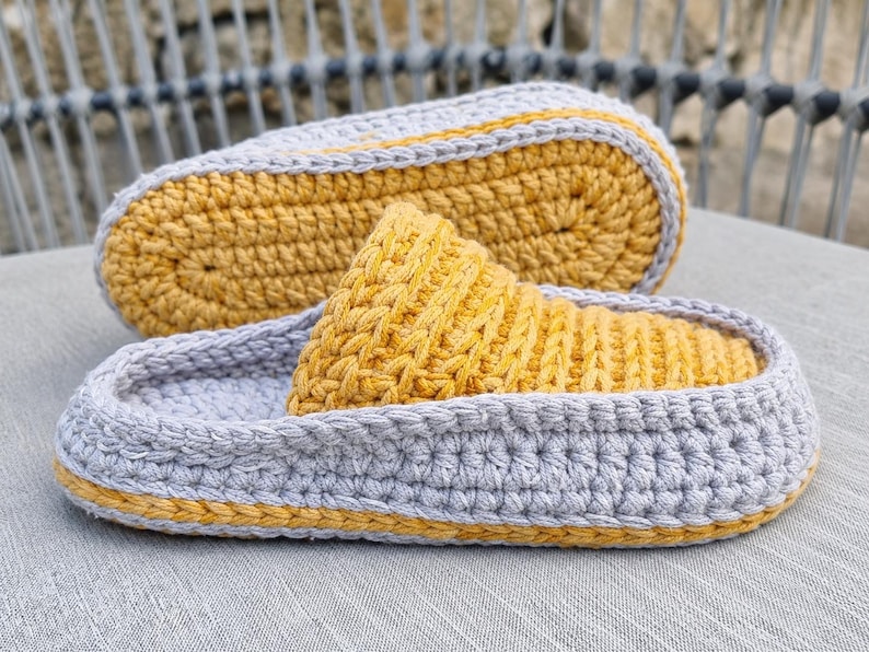 May include: Handmade crochet slippers in gray and yellow. The slippers feature a gray base with a yellow sole and a yellow top. The crochet stitch is visible, adding texture. The slippers are designed for indoor use and provide comfort and warmth.