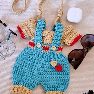 May include: A handmade crochet baby outfit in blue, tan, and red. The outfit includes overalls with wooden buttons and a matching top. A tan strap is attached for carrying. Two heart appliqués are on the front.