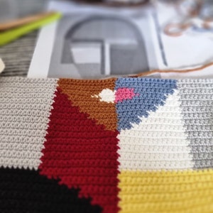May include: Close-up of a colorful, geometric crochet project in progress. The design features blocks of red, brown, blue, yellow, gray, and black. A basket of yarn and a pattern are blurred in the background, suggesting a craft project.
