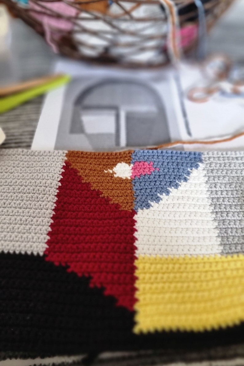 May include: Close-up of a colorful, geometric crochet project in progress. The design features blocks of red, brown, blue, yellow, gray, and black. A basket of yarn and a pattern are blurred in the background, suggesting a craft project.