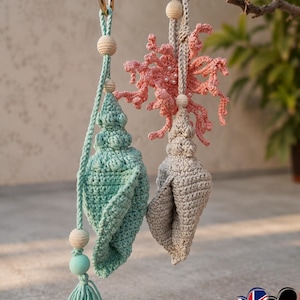 Op de afbeelding: Twee handgemaakte gehaakte ornamenten die aan een tak hangen. Eén is zeegroen met een kwast, de andere is grijs. Beide hebben houten kralen en goudkleurige ringen. Het derde ornament is koraalkleurig en lijkt op koraal.