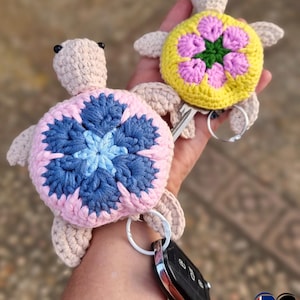 May include: Two handmade crochet turtle keychains. One has a pink and blue flower design, the other a yellow and pink flower design. Both have beige bodies and are attached to key rings. The keychains are held in a hand.