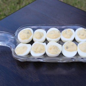 May include: A clear glass serving dish shaped like a bottle, filled with deviled eggs. The eggs are arranged in a row and are topped with a yellow filling.