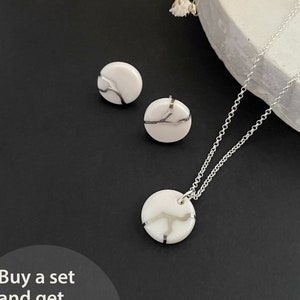 May include: A set of white ceramic earrings and a necklace with a silver chain. Each piece features a delicate silver design on a white background. The text "Buy a set and get 10% OFF * the discount is included in the price" is displayed below the jewelry.