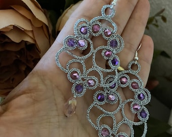 May include: A pair of silver earrings with a delicate, intricate design featuring a swirling pattern and purple and pink beads. The earrings have a dangling drop design with clear beads.