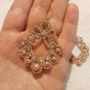 Può includere: Un paio di orecchini dorati con un delicato design all'uncinetto e perle rosa. Gli orecchini hanno la forma di un fiocco con un piccolo cristallo al centro.