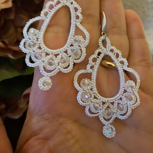 Può includere: Un paio di orecchini in crochet bianco con dettagli in cristallo. Gli orecchini hanno una forma a goccia e un design delicato e intricato.