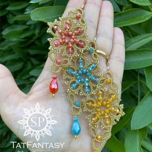 Può includere: Orecchini dorati fatti a mano con intricati disegni floreali. Ogni orecchino presenta perline colorate rosse, blu e gialle, con pendenti a goccia abbinati. Gli orecchini sono tenuti su uno sfondo di fogliame verde.