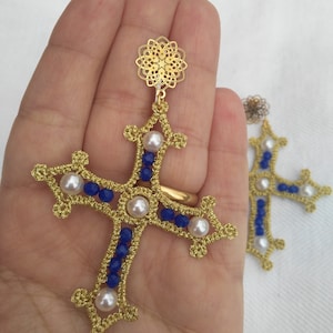 Può includere: Orecchini a forma di croce dorati con perline blu e bianche. La croce è realizzata con un lavoro di filigrana intricato e presenta un delicato motivo floreale nella parte superiore.