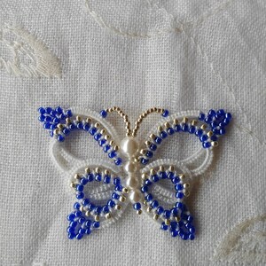 May include: A white and blue beaded butterfly with pearl accents. The butterfly is made with white thread and blue and gold beads.