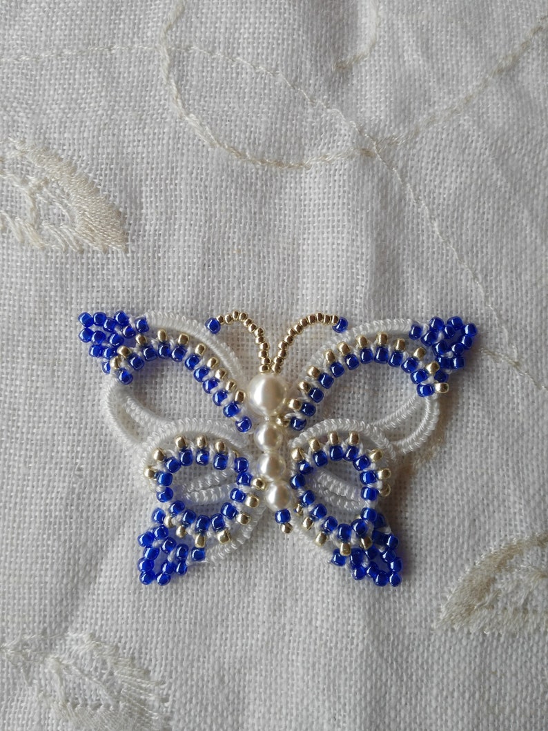 May include: A white and blue beaded butterfly with pearl accents. The butterfly is made with white thread and blue and gold beads.