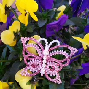 May include: A pink and white beaded butterfly brooch with pearl accents. The brooch is sitting on a bed of purple and yellow flowers.