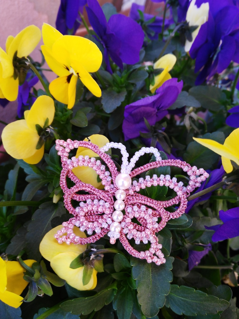 May include: A pink and white beaded butterfly brooch with pearl accents. The brooch is sitting on a bed of purple and yellow flowers.