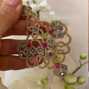 Può includere: Orecchino in pizzo dorato con cristalli blu e rosa. L'orecchino è delicato e intricato, con un design floreale.