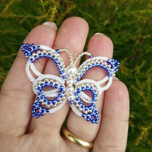 May include: A white and blue beaded butterfly brooch with a pearl accent. The butterfly is made with small seed beads and has a delicate, intricate design.