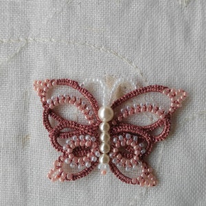 May include: A pink and white beaded butterfly with pearl accents. The butterfly is made with a crocheted design and is a delicate piece of jewelry.