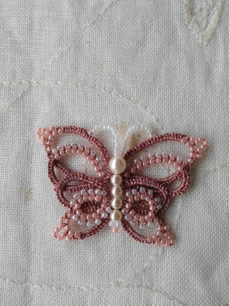 May include: A pink and white beaded butterfly with pearl accents. The butterfly is made with a crocheted design and is a delicate piece of jewelry.