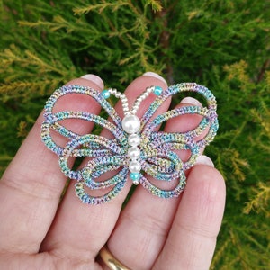 May include: A colorful butterfly-shaped brooch made of wire and beads. The butterfly has a pearl in the center and is decorated with iridescent blue, green, and purple beads.