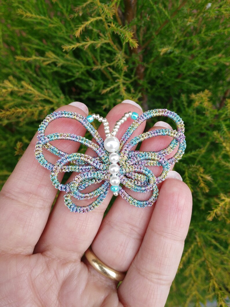 May include: A colorful butterfly-shaped brooch made of wire and beads. The butterfly has a pearl in the center and is decorated with iridescent blue, green, and purple beads.
