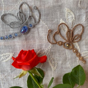 Può includere: Due spille a forma di libellula in perle, una argentata con perle blu e una dorata con perle trasparenti, su uno sfondo di tessuto bianco con una rosa rossa.
