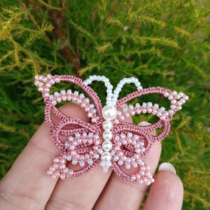 May include: A pink and white beaded butterfly with a pearl in the center. The butterfly is made of thread and beads and is a delicate piece of jewelry.