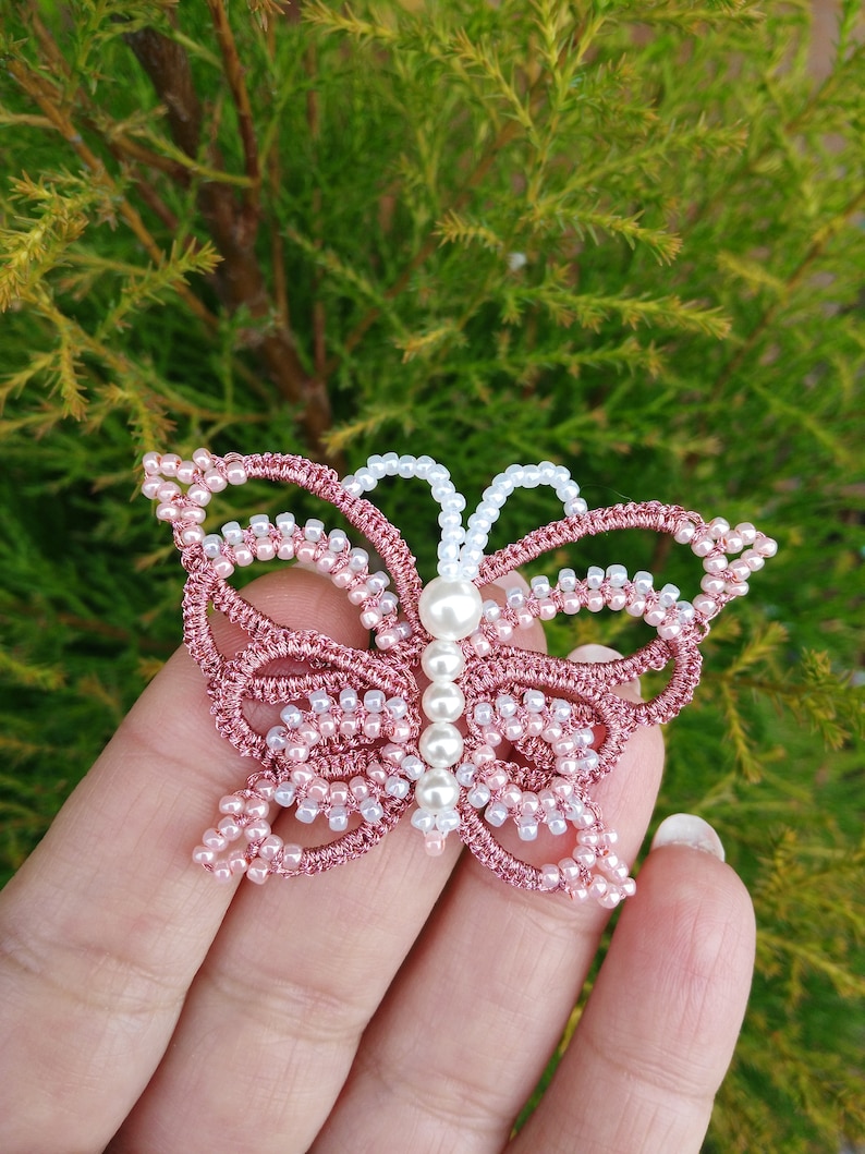 May include: A pink and white beaded butterfly with a pearl in the center. The butterfly is made of thread and beads and is a delicate piece of jewelry.