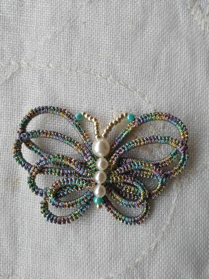 May include: A colorful butterfly brooch made of woven thread with a pearl and turquoise bead accents.