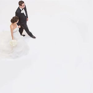 May include: A bride and groom walking towards the camera, the bride is wearing a white wedding dress with a long train and holding a bouquet of white flowers, the groom is wearing a black suit with a white shirt and a black bow tie.